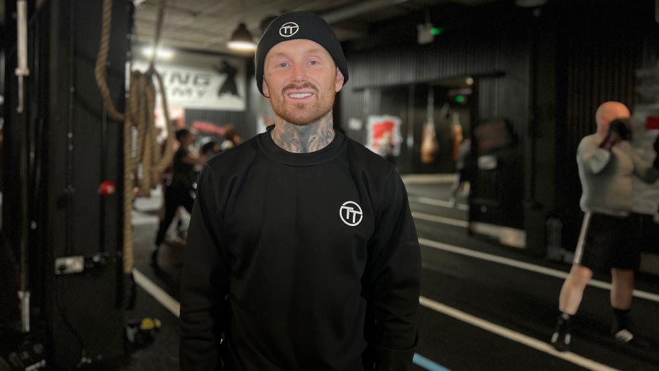 Cameron smiles at the camera in the boxing gym. Behind him people are sparring. Camera is wearing a black jumper and knitted hat with the gym's TT logo on them. He has a stubbly ginger beard and tattoos on his neck.